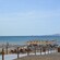 A public beach facing the Aegean Sea, featuring a wooden walkway leading to an area with a large number of sunbeds and umbrellas